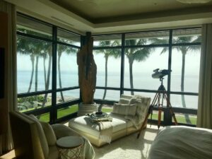 An interior space shows a chaise lounge, telescope on a tripod, and large floor-to-ceiling windows framing palm trees and the ocean.