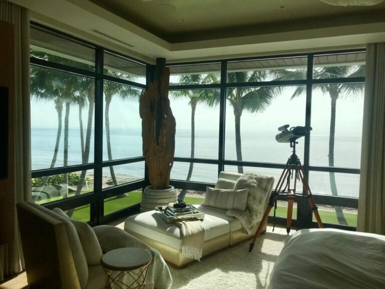 An interior space shows a chaise lounge, telescope on a tripod, and large floor-to-ceiling windows framing palm trees and the ocean.