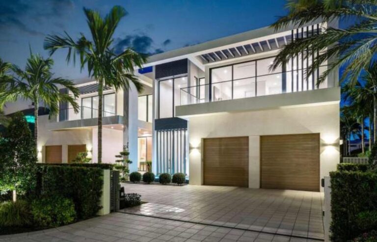 A modern two-story house with large glass windows, two wooden garage doors, and palm trees in the front driveway.