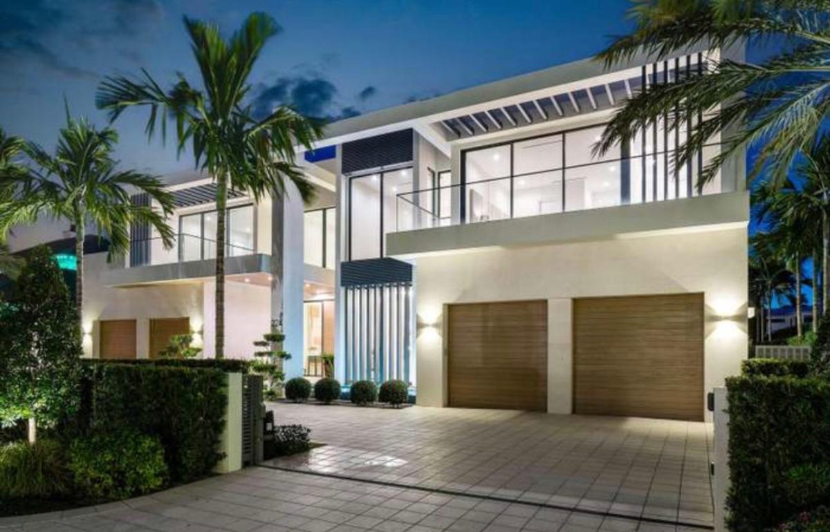 A modern two-story house with large glass windows, two wooden garage doors, and palm trees in the front driveway.