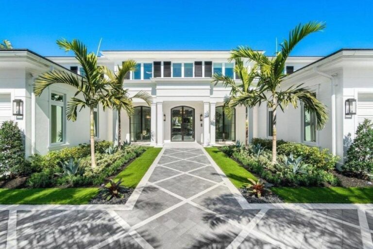 A patterned paved walkway leads to double glass doors at the entrance of a white modern house flanked by palm trees and landscaped beds.