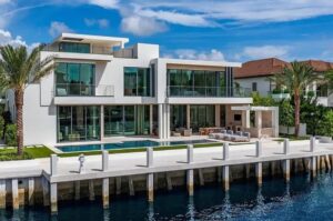 A modern multi-story waterfront house with large glass windows, a swimming pool, and a dock.