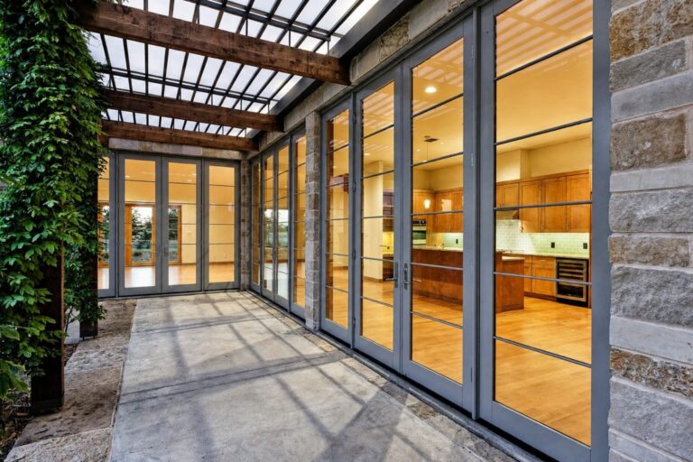 A covered patio with a wooden pergola faces a row of tall glass doors revealing a warmly lit kitchen with wooden cabinets.