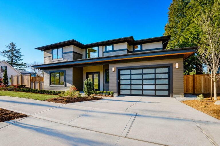 A modern two-story house with a glass-panel garage door and landscaped front yard is shown under a clear blue sky.