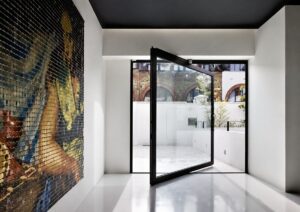 A minimalist interior featuring a large black-framed pivoting glass door open to an outdoor terrace and a tiled mosaic artwork on the left wall.